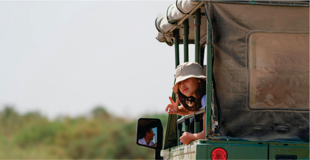 Guide visible on a side mirror and a young girl sticking her head out of an Amboseli safari vehicle rental with a blurred natural background