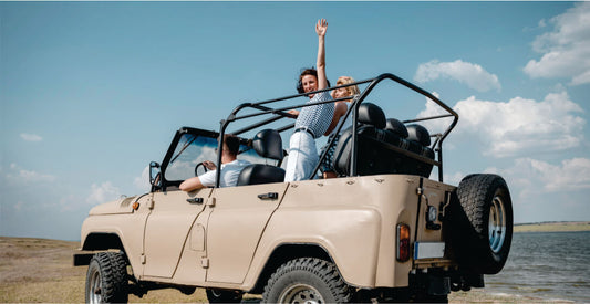 Two people in an open-top beige jeep by a body of water with a clear blue sky.