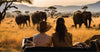 Two people sitting in a safari vehicle on Masai Mara Safari 3 days game drive package in Kenya, observing elephants in a grassy savanna at sunset.
