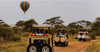Three safari vehicles on a dirt road with a hot air balloon in the sky.