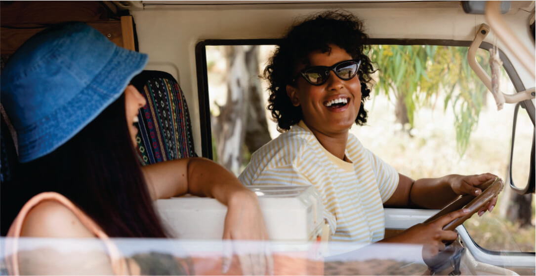 Two people inside a vehicle, one driving and smiling, with a scenic background.